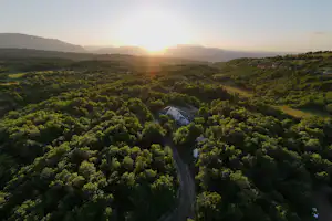 Blick auf ein Tal mit dichtem Wald und einem schmalen Weg, im Hintergrund geht die Sonne auf.