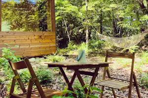Holzbank und Tisch unter einem kleinen Unterstand am Campingplatz, umgeben von dichtem Grün und Bäumen.