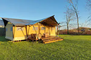 Zelt mit Veranda auf grünem Campingplatz, Holzpodest und Sitzbank, im Hintergrund Zaun und Bäume.