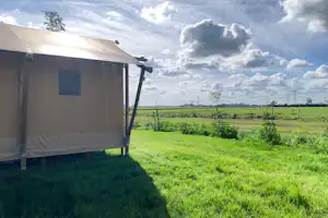 Campingzelt mit Fenster und Markise am Rand eines grünen Feldes, im Hintergrund weite Wiesen und Wolkenhimmel.