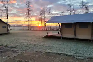 Campingzelt mit Holzpodest und Markise am Stellplatz, im Hintergrund Bäume und ein Sonnenaufgang am Horizont.