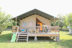Zeltbungalow mit Veranda, Holztreppe und Kinderzaun; im Vordergrund ein grütsitzender Spielbereich im Garten.