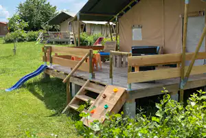 Spielbereich auf einer Holzplattform mit Rutsche, orangefarbenen Stühlen und Zeltüberdachung im grünen Garten.