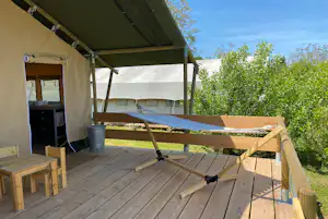 Glamping-Zelt mit Holzterrasse, Markise und Liege, im Hintergrund weitere Zelte und grüner Garten mit Büschen.
