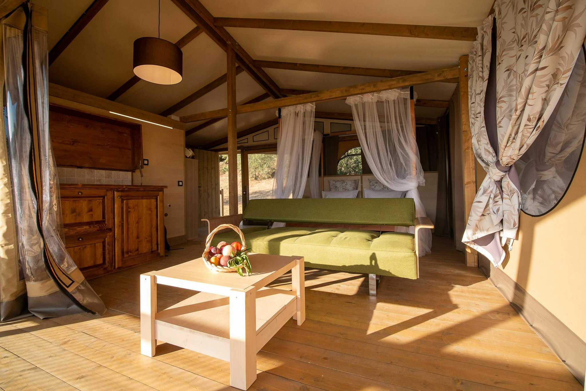 Innenansicht eines Wohnwagens mit Doppelbett, Sitzbank und Holztisch, Vorhängen und warmem Sonnenlicht.