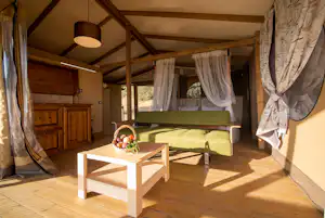 Innenansicht eines Wohnwagens mit Doppelbett, Sitzbank und Holztisch, Vorhängen und warmem Sonnenlicht.