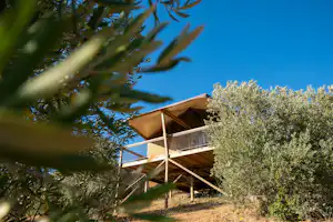 Camping-Unterkunft auf Stelzen mit Terrasse, umgeben von Bäumen und Sträuchern vor blauem Himmel.