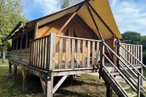 Gelbes Zelt auf Stelzen mit Holzgeländer und Treppe, umgeben von Bäumen und Rasen auf dem Campingplatz.