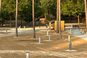 Campingplatz mit Stellplätzen, Betonflächen und Stromsäulen, umgeben von hohen Bäumen und einem Zaun im Hintergrund.
