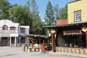 Überdachter Bereich mit Grill und Theke am Campingplatz, daneben Sitzbänke und Tische vor Holzhäusern und Bäumen.