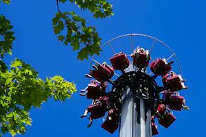 Achterbahn mit roten Sitzen und Sicherheitsbügeln, aufgenommen von unten vor blauem Himmel und grünen Baumkronen.