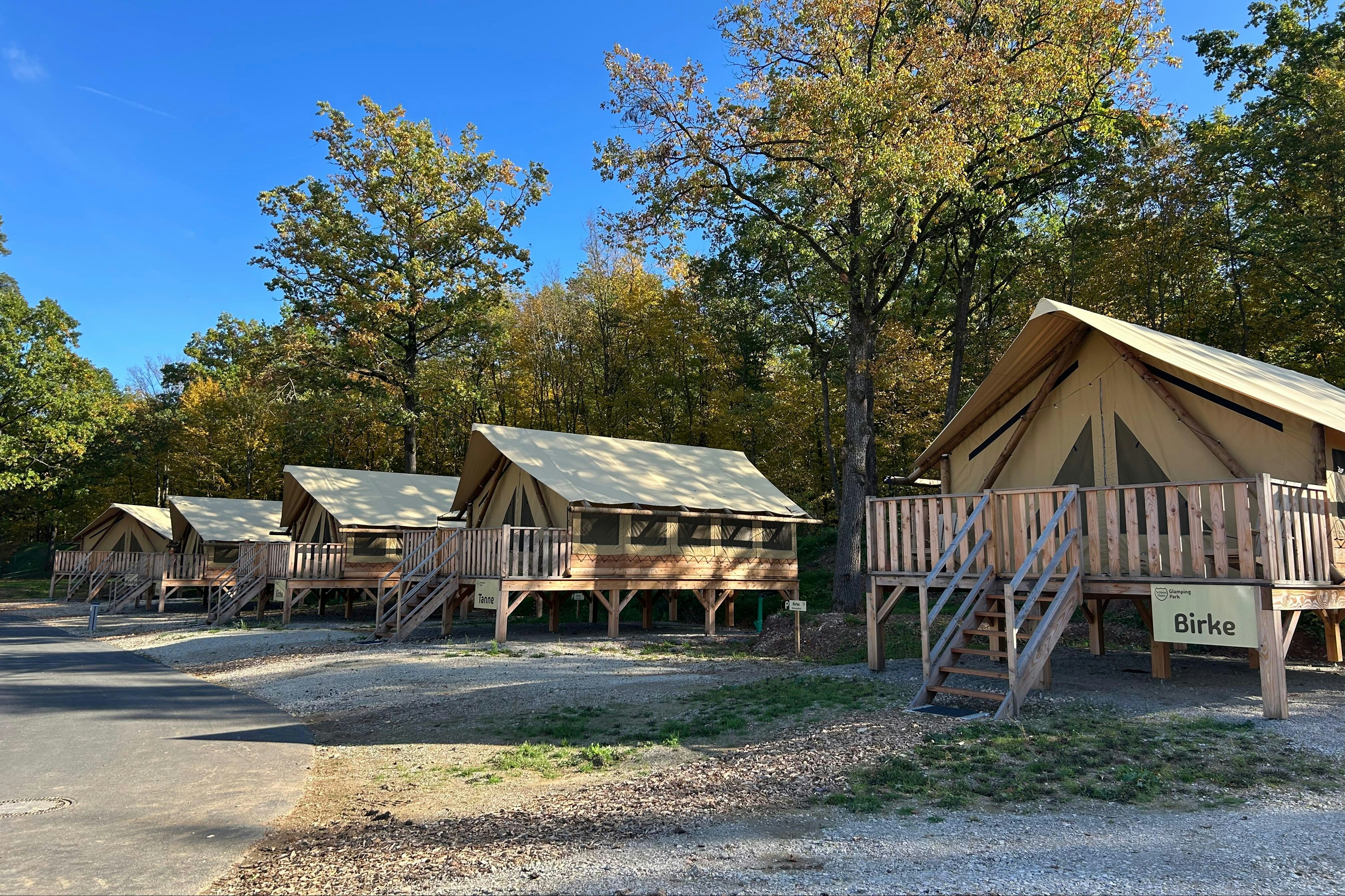 Holzhütten und Zelte auf einem Campingplatz, mit Holzterrassen und Treppen, umgeben von Bäumen und einem Weg.
