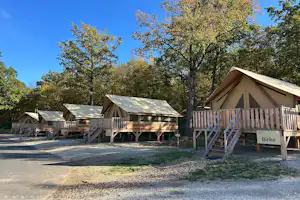 Holzhütten und Zelte auf einem Campingplatz, mit Holzterrassen und Treppen, umgeben von Bäumen und einem Weg.