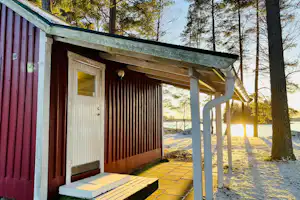 Holzhütte mit Veranda, Holzjalousien und weißen Rohren, davor gepflasterter Weg und Bäume im Hintergrund.