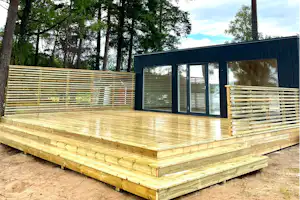 Holzterrasse mit Zaun aus Holzlatten und großen Glastüren an einem modernen Gebäude im Wald.