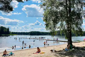 Seeufer mit Sandstrand, Steg und Stand-up-Paddle-Boardern; im Hintergrund Wald und viele Wolken am Himmel.