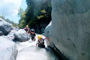 Eine Familie klettert und watet im Fluss zwischen großen Felsen, im Hintergrund Bäume und Felswand.