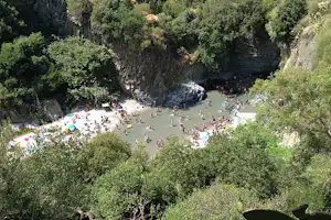 Fluss in einem grünen Tal mit vielen Badegästen am Ufer, umgeben von Bäumen und Felsen.