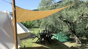 Zelt mit Sonnensegel, daneben Tisch und Stühle auf dem Campingplatz im Grünen, Hecke und Bäume im Hintergrund.