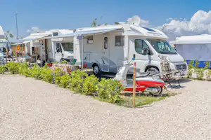 Weißes Wohnmobil auf einem Stellplatz mit Markise, davor Tisch und Stühle, umgeben von Sträuchern und Kiesweg.