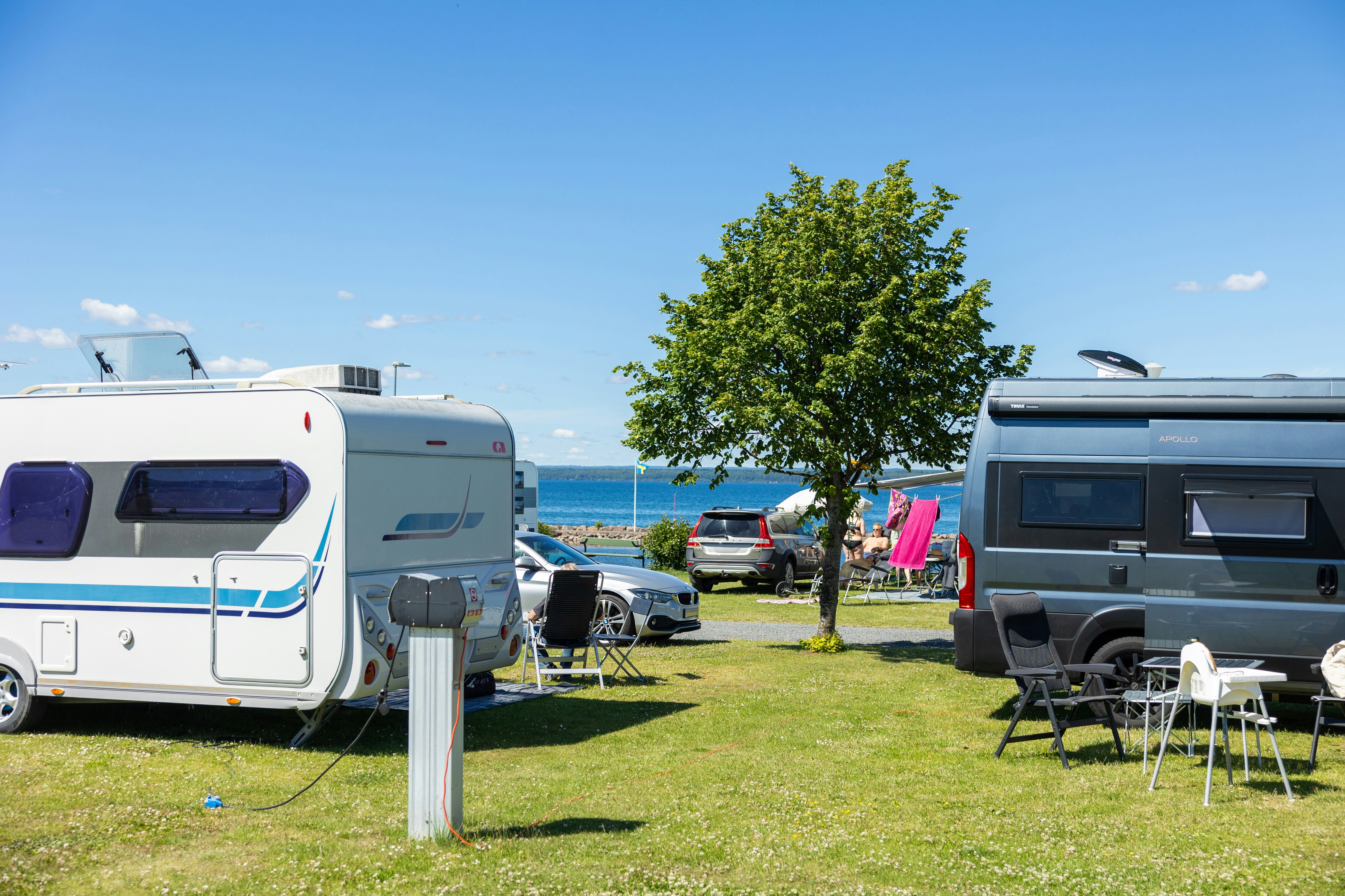 Wohnmobile auf einem Campingplatz, mit Sitzgarnitur und Tisch am Gras, im Hintergrund ein See und ein Baum.