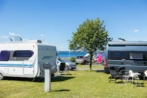 Wohnmobile auf einem Campingplatz, mit Sitzgarnitur und Tisch am Gras, im Hintergrund ein See und ein Baum.