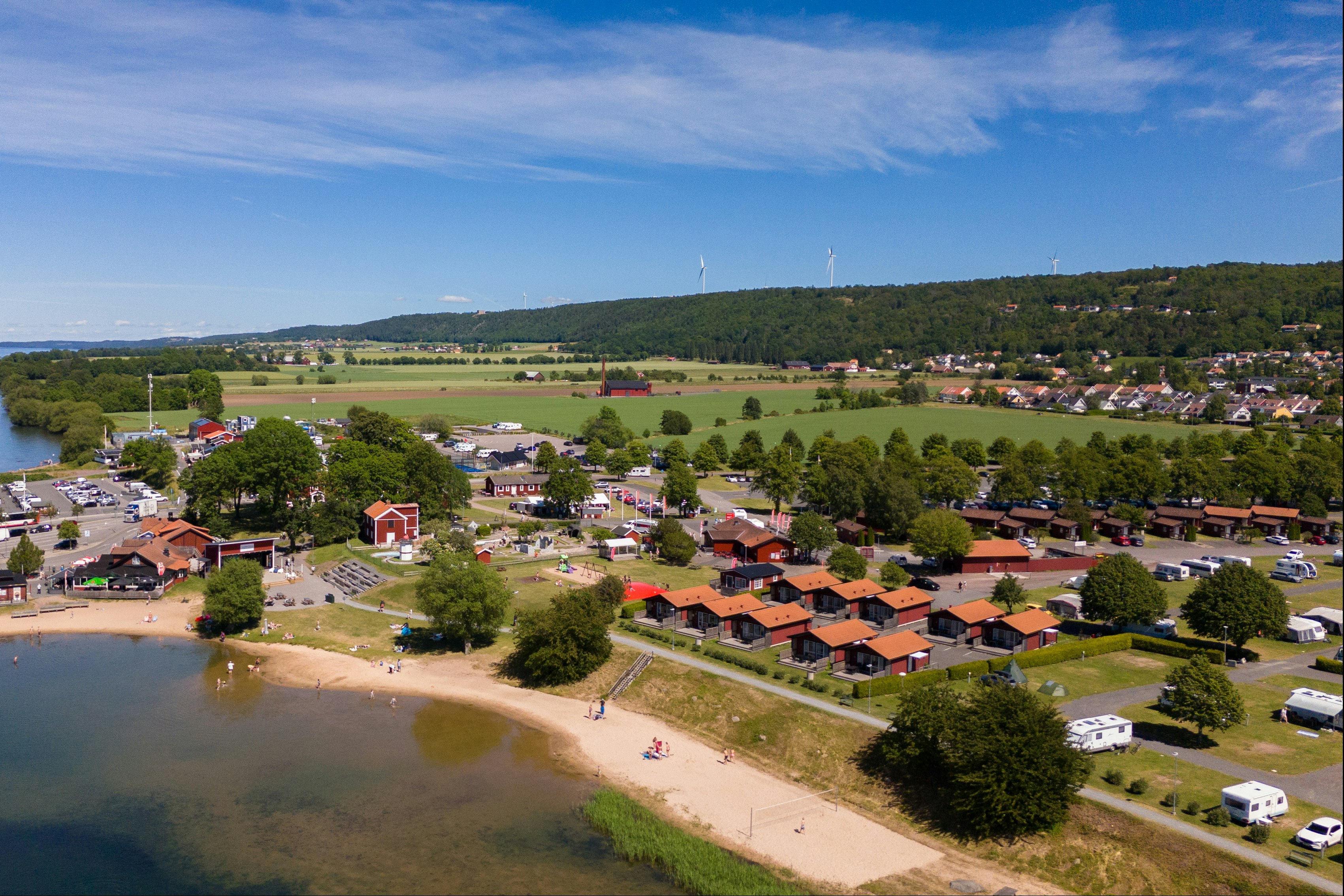 Luftaufnahme eines Campingplatzes am See mit Uferpromenade, Stellplätzen, Wohnwagen und Häusern am Hang.