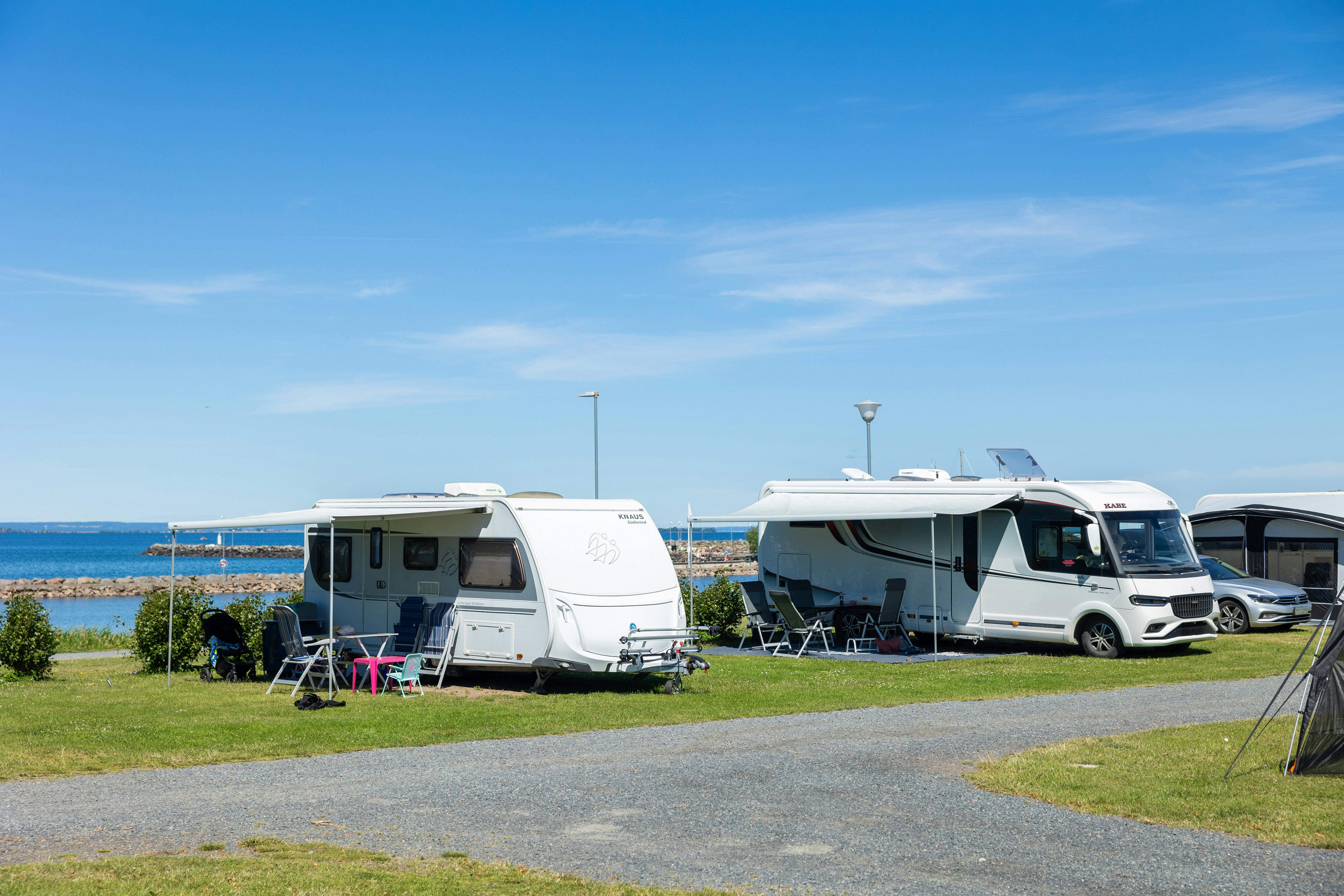 Wohnmobile auf einem Campingplatz mit Rasenfläche, Markise und Stühlen; im Hintergrund Meer und blauer Himmel.