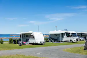 Wohnmobile auf einem Campingplatz mit Rasenfläche, Markise und Stühlen; im Hintergrund Meer und blauer Himmel.