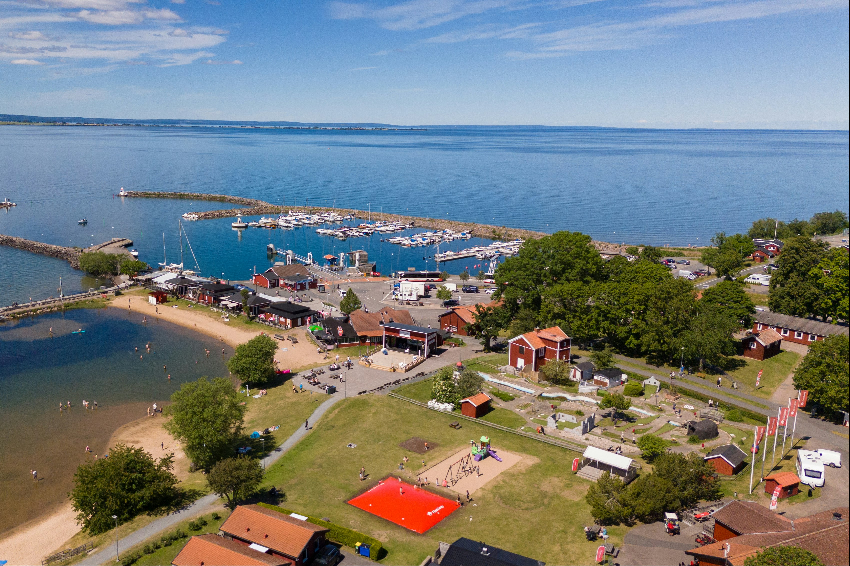 Blick auf einen Campingplatz am See mit Stegen, Booten, Uferweg und Rasenflächen, daneben Gebäude und Bäume.