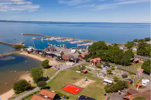 Blick auf einen Campingplatz am See mit Stegen, Booten, Uferweg und Rasenflächen, daneben Gebäude und Bäume.