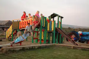 Kinder spielen auf einer bunten Rutsche am Spielplatz, im Hintergrund Wohnwagen und Landschaft mit Feldern.