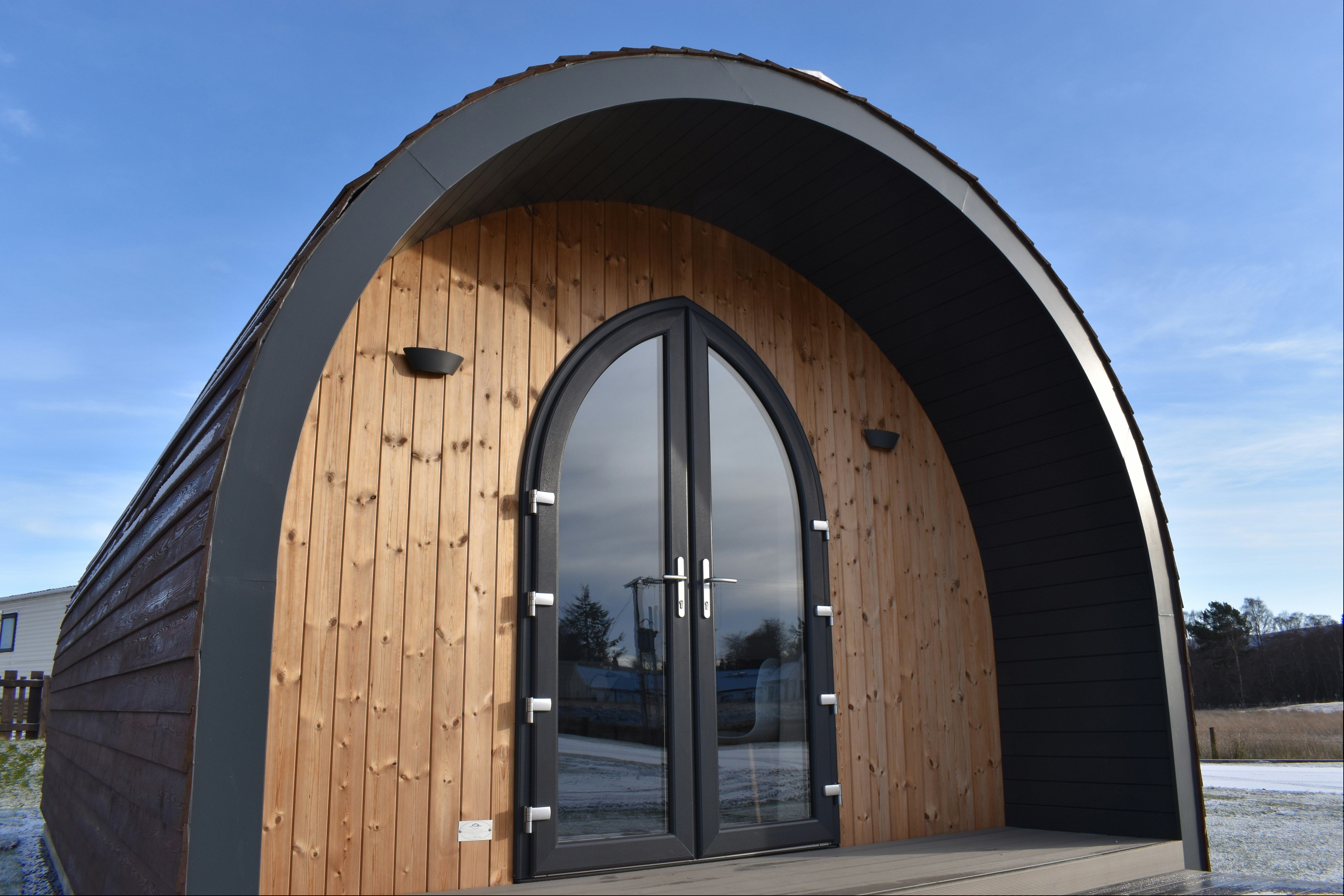 Holz-Iglu-Zelt mit schwarzem Eingang und Glastür, auf einem gepflasterten Platz, im Hintergrund Gebäude und Himmel.