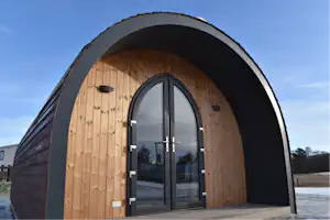 Holz-Iglu-Zelt mit schwarzem Eingang und Glastür, auf einem gepflasterten Platz, im Hintergrund Gebäude und Himmel.