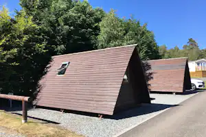 Holz-Hütte mit Satteldach auf Kiesstellplatz, daneben weitere Hütte und Auto, umgeben von hohen Bäumen.