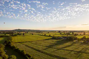 Weite Landschaft mit grünen Feldern, Hecken und einem kleinen Dorf in der Ferne unter wolkigem Himmel.