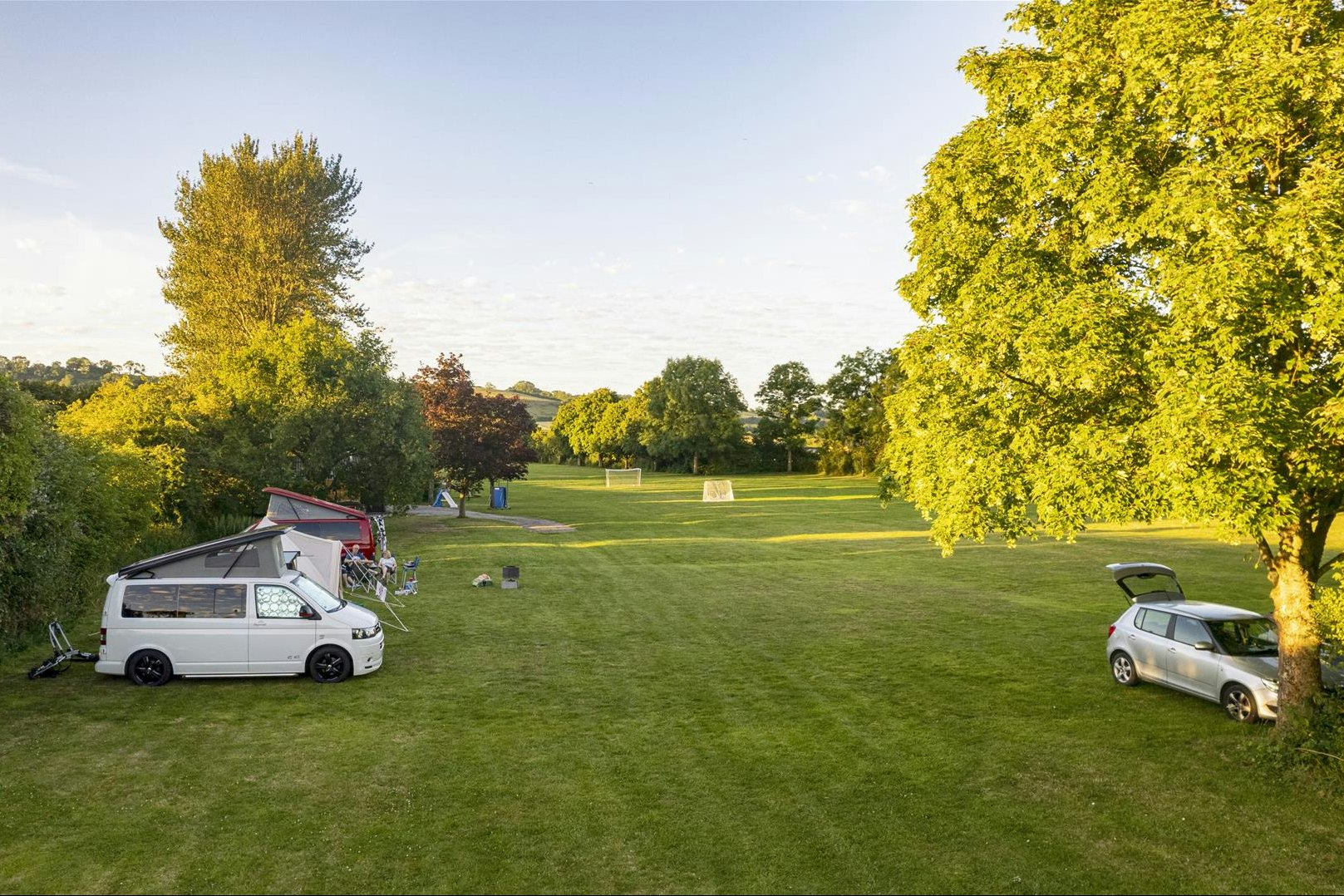 Wohnmobil auf einem Stellplatz auf grünem Campinggelände, daneben ein Auto, Bäume und Rasenfläche im Hintergrund.