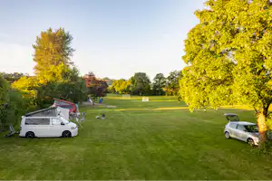 Wohnmobil auf einem Stellplatz auf grünem Campinggelände, daneben ein Auto, Bäume und Rasenfläche im Hintergrund.