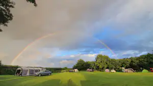Campingwiese mit Wohnwagen und Zelt, im Hintergrund Bäume und ein Regenbogen am Himmel.