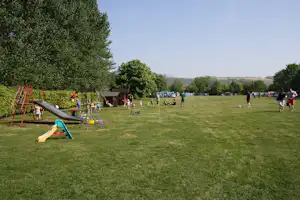 Große Campingwiese mit Spielplatz, Schaukel und Rutsche, daneben Bäume und mehrere Personen auf dem Rasen.