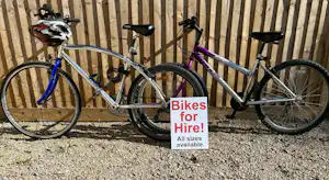 Zwei Fahrräder vor einer Holzfassade, mit Schild „Bikes for Hire!“ und Hinweis „All sizes available“.