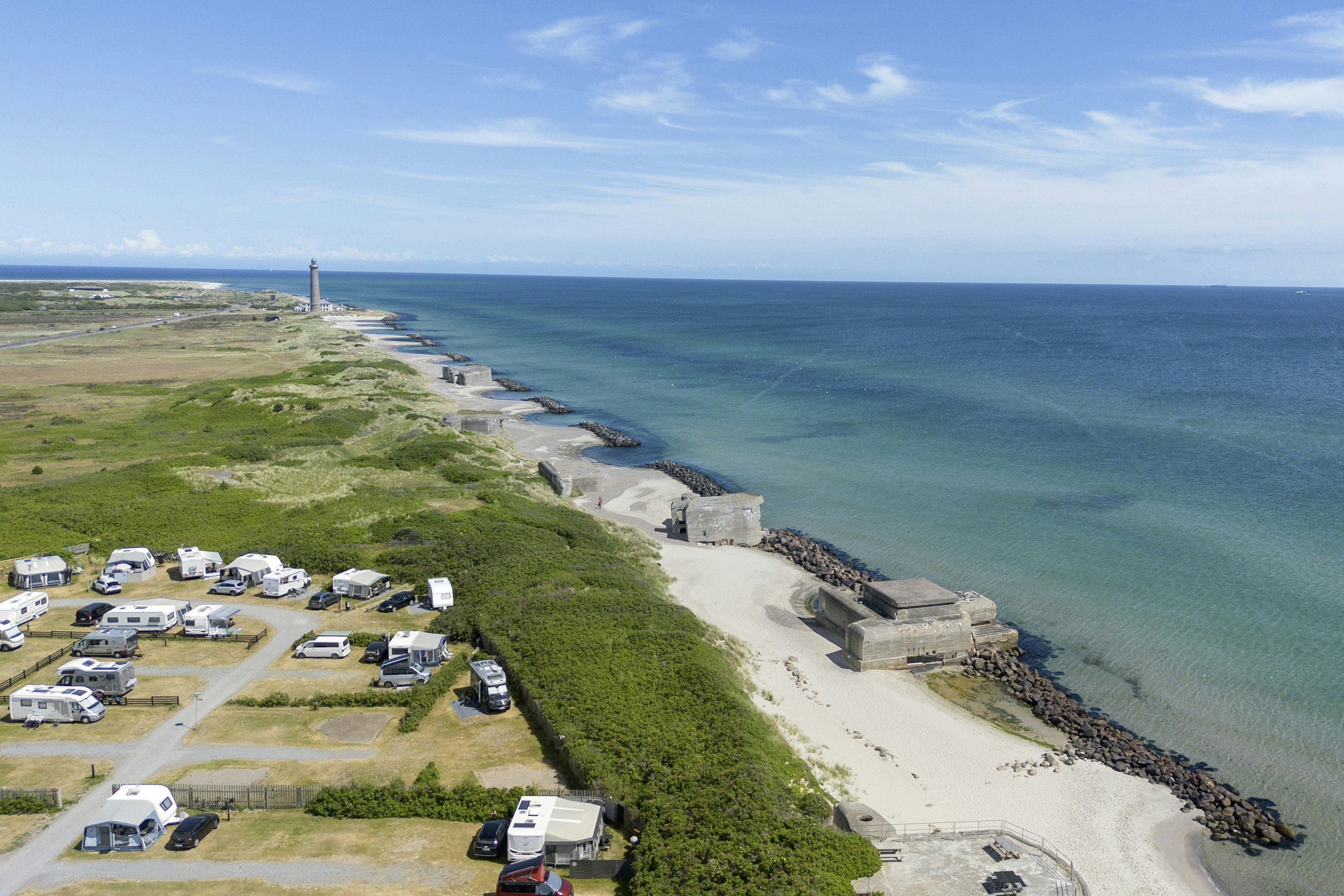 Campingplatz mit Wohnwagen und Stellplätzen am Küstenweg, Blick auf das Meer und den Strandabschnitt mit Wellenbrechern.