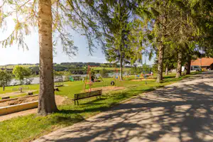 Spielplatz mit rotem Schaukelsystem und Sandfläche am Campingplatz, Bänke, Bäume und Blick auf den See.