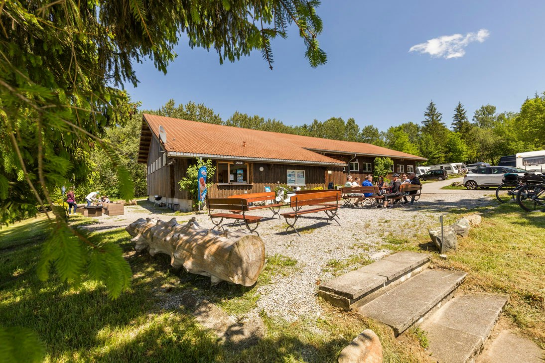 Holzgebäude mit Terrasse und Sitzbänken, davor Kiesweg und große Baumstämme, rechts geparkte Autos und Fahrräder.