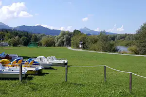 Ruderboote am Steg neben einer grünen Wiese, im Hintergrund See und bewaldete Hügel.