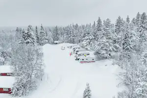 Verschneite Campinganlage mit Wohnwagen und kleinen Gebäuden, umgeben von Tannen und verschneiten Wegen im Winter.