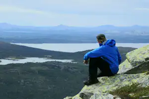 Wanderer sitzt auf einem Felsen und blickt über ein weites Tal mit See, Wäldern und Bergen in der Ferne.