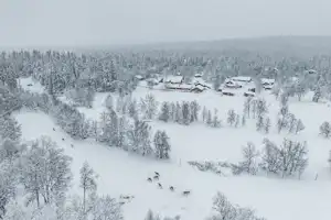 Winterlicher Campingplatz im Schnee mit mehreren Hütten und Gebäuden, umgeben von verschneiten Bäumen und Wiesen.