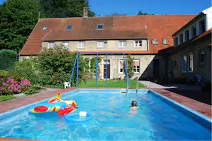 Schwimmbecken auf dem Campingplatz mit aufblasbarem Spielzeug, Schaukelgestell und Wohngebäude im Hintergrund.