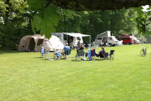 Campingplatz mit mehreren Zelten und Wohnwagen auf einer grünen Wiese, daneben Campingstühle und Tische unter Bäumen.